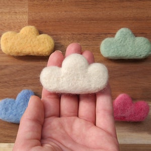 Może przedstawiać: Pięć filcowych kształtów chmur w różnych kolorach. Dłoń trzyma biały kształt chmury w środku obrazu. Pozostałe chmury są żółte, zielone, niebieskie i różowe.