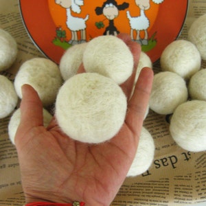 May include: A close-up of a hand holding a white wool dryer ball. There are several other white wool dryer balls scattered around the hand on a brown surface.
