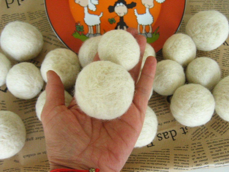 May include: A close-up of a hand holding a white wool dryer ball. There are several other white wool dryer balls scattered around the hand on a brown surface.