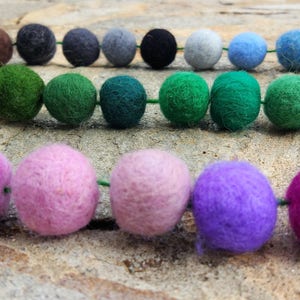 May include: A garland of colorful felt balls in shades of green, blue, purple, pink, and brown. The balls are strung together with green thread.