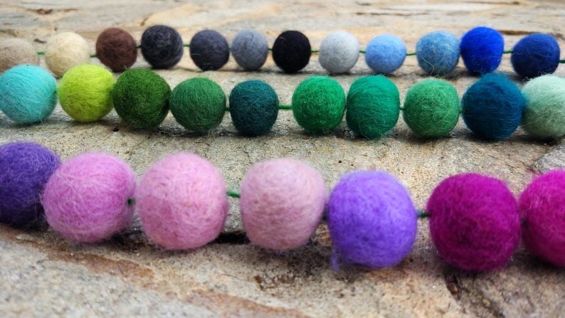 May include: A garland of colorful felt balls in shades of green, blue, purple, pink, and brown. The balls are strung together with green thread.
