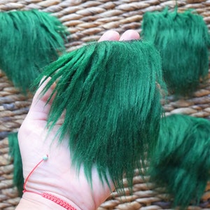 May include: Close-up of several pieces of bright green faux fur with a long, shaggy texture. The fur is held in a hand against a woven background. The pieces are cut into irregular shapes, possibly for crafting or costume design.