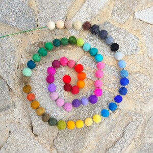 Pode incluir: Uma guirlanda colorida de bolas de feltro em um padr&atilde;o de arco-&iacute;ris. A guirlanda est&aacute; disposta em espiral sobre um fundo de pedra.