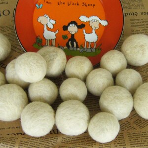 May include: A close-up of a group of white wool dryer balls. The balls are arranged in a circle on a brown and white patterned surface. A plate with a cartoon image of three sheep is visible in the background.