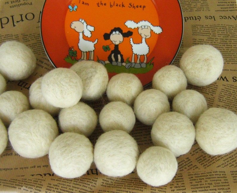 May include: A close-up of a group of white wool dryer balls. The balls are arranged in a circle on a brown and white patterned surface. A plate with a cartoon image of three sheep is visible in the background.