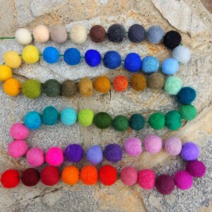 May include: A colorful garland made of felt balls in various shades of red, orange, yellow, green, blue, purple, and brown. The garland is strung on a green wire and is arranged in a rainbow pattern.