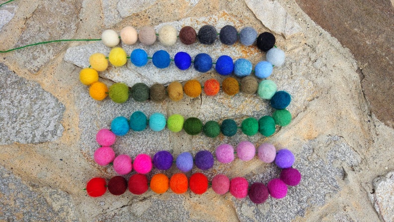 May include: A colorful garland made of felt balls in various shades of red, orange, yellow, green, blue, purple, and brown. The garland is strung on a green wire and is arranged in a rainbow pattern.