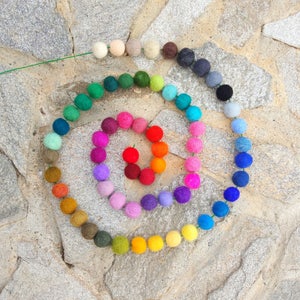 May include: A colorful garland of felt balls in a rainbow pattern. The garland is arranged in a spiral shape on a stone surface.