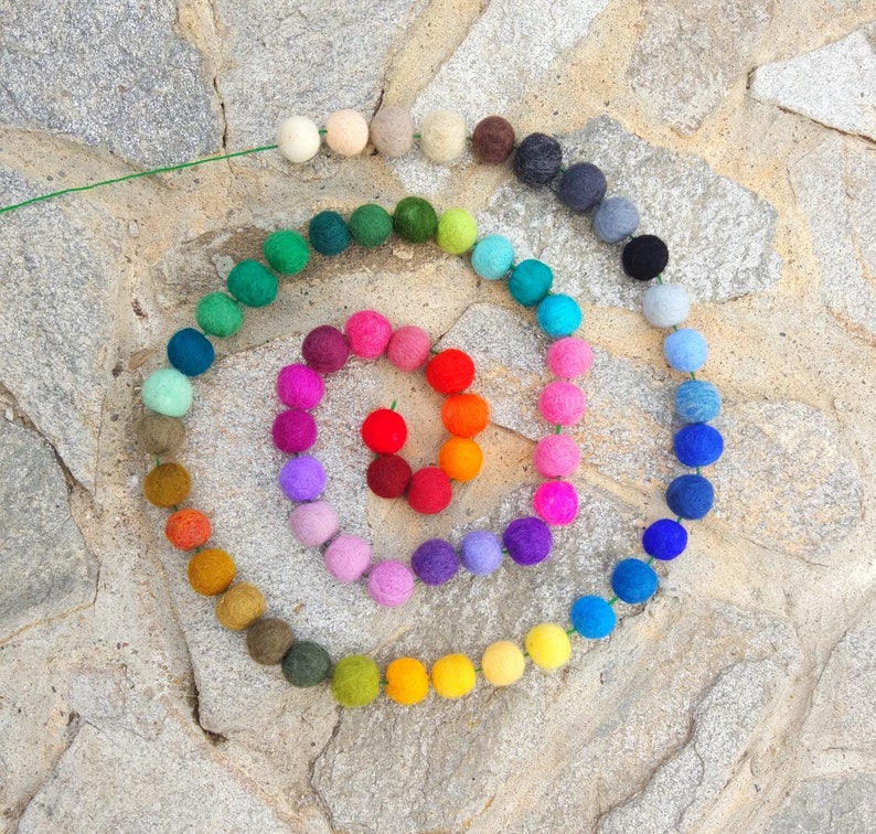 May include: A colorful garland of felt balls in a rainbow pattern. The garland is arranged in a spiral shape on a stone surface.
