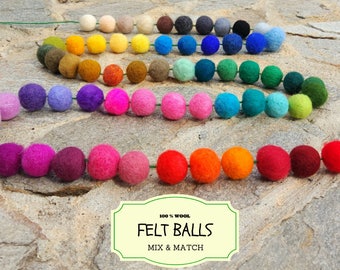 May include: A colorful garland of 100% wool felt balls. The garland is strung on a green wire and features a variety of colors, including red, orange, yellow, green, blue, purple, and pink. The felt balls are all approximately the same size and are arranged in a rainbow pattern. The garland is displayed on a stone surface. 100% Wool Felt Balls Mix & Match