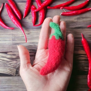 May include: A hand holding a red felt chili pepper with a green felt stem. The chili pepper is a handmade craft item.