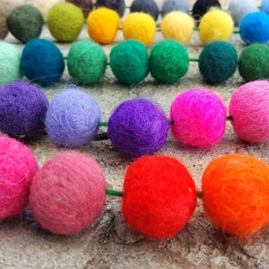 May include: A colorful garland of felt balls in various shades of green, blue, purple, pink, red, and orange. The balls are strung together on a green string.