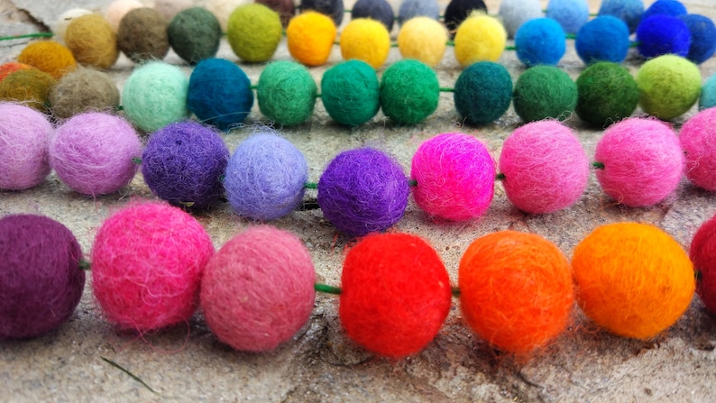 May include: A colorful garland of felt balls in various shades of green, blue, purple, pink, red, and orange. The balls are strung together on a green string.