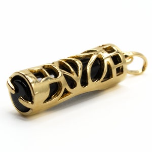 May include: A gold-colored pendant with a cylindrical shape. The pendant features an openwork design with intricate cutouts, revealing a black interior. The ends of the pendant are capped with black, rounded stones, and it has a small loop for a chain.