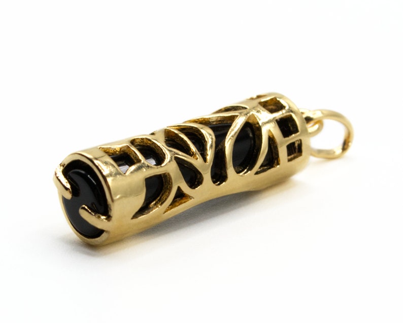 May include: A gold-colored pendant with a cylindrical shape. The pendant features an openwork design with intricate cutouts, revealing a black interior. The ends of the pendant are capped with black, rounded stones, and it has a small loop for a chain.