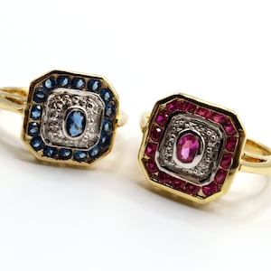 May include: Two gold rings with square settings. The ring on the left has a blue gemstone surrounded by smaller blue gemstones and white diamonds. The ring on the right has a red gemstone surrounded by smaller red gemstones and white diamonds.