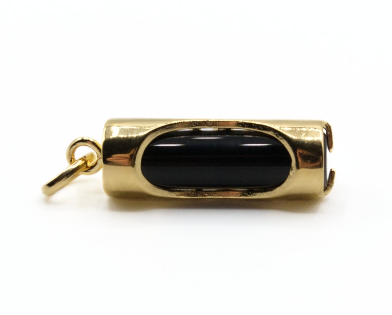 May include: A gold-colored pendant with a black, cylindrical stone set in the center. The pendant has a rounded rectangular shape with an oval opening on each side, and a small loop for attaching to a chain.