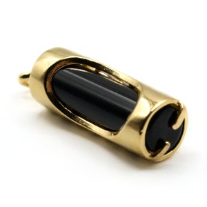 May include: A gold-colored pendant with a cylindrical shape. The pendant features a black, oval-shaped stone set within a gold frame. The design includes cut-out sections on the sides and a small loop for a chain.