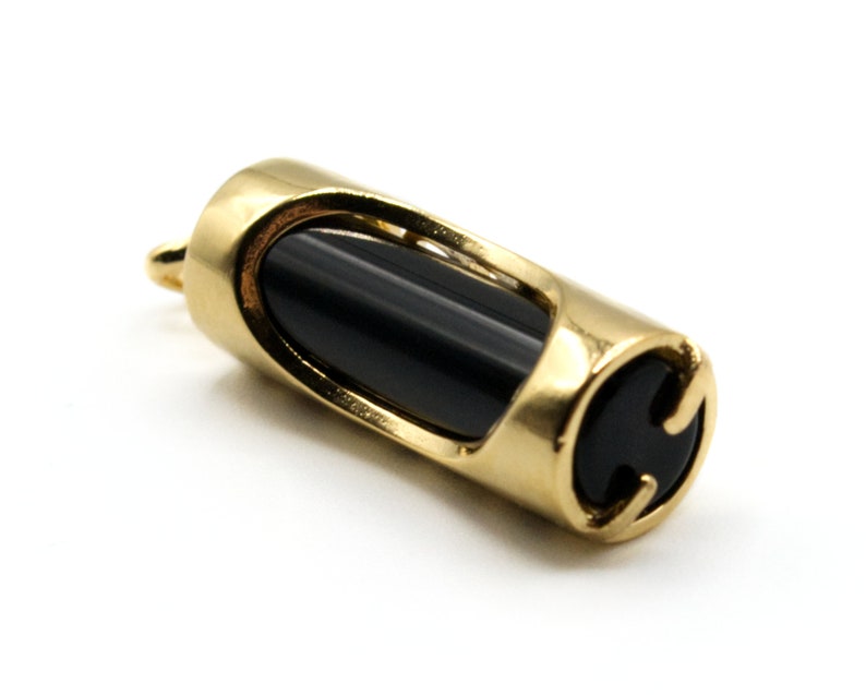 May include: A gold-colored pendant with a cylindrical shape. The pendant features a black, oval-shaped stone set within a gold frame. The design includes cut-out sections on the sides and a small loop for a chain.