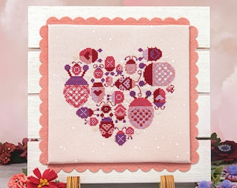 Peut inclure: Une broderie au point de croix représentant un cœur composé d'une collection de coccinelles roses et violettes. La broderie est encadrée dans un cadre en bois blanc avec une bordure rose festonnée.