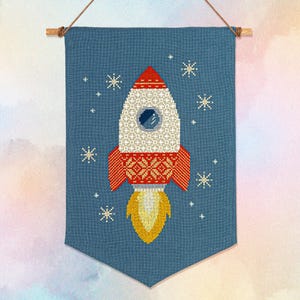 Op de afbeelding: Een blauwe banner met een kruissteekontwerp van een raket. De raket is wit met rode accenten en een gele vlam. Witte stervormige ontwerpen zijn rond de raket verspreid.