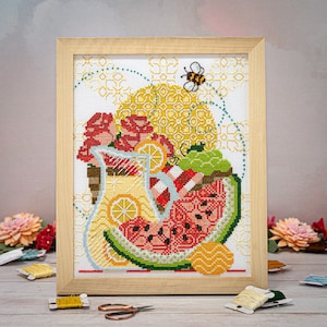 Può includere: Ricamo a punto croce che rappresenta una brocca piena di limoni, anguria e fiori. La brocca è circondata da un motivo geometrico giallo e bianco. Un'ape vola sopra la brocca.