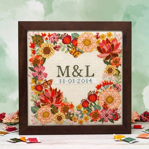 May include: A framed cross-stitch embroidery featuring a floral wreath design in shades of red, orange, yellow, and pink. The centre of the wreath displays the initials "M&L" and the date "XX-0X-20XX".