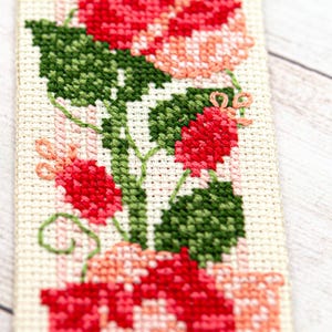 Pode incluir: Marcador de livros de ponto cruz feito &agrave; m&atilde;o com um design floral. O marcador apresenta flores vermelhas e rosa com folhas verdes, todas costuradas em tecido creme. O arranjo floral &eacute; detalhado e vibrante.