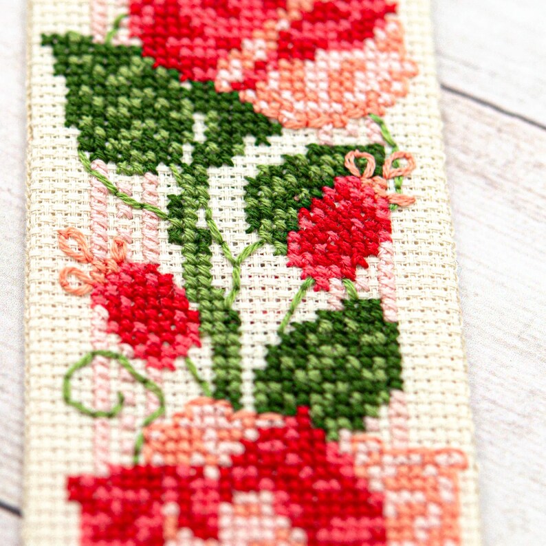 Pode incluir: Marcador de livros de ponto cruz feito &agrave; m&atilde;o com um design floral. O marcador apresenta flores vermelhas e rosa com folhas verdes, todas costuradas em tecido creme. O arranjo floral &eacute; detalhado e vibrante.