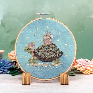 Könnte beinhalten: Ein blauer und weißer Stickrahmen mit einem Kreuzstich-Design einer Schildkröte, die ein Geschenk trägt. Die Schildkröte ist mit grünen, braunen und rosa Details verziert. Der Hintergrund ist hellblau mit weißen Sternen.