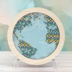 May include: A round, cross-stitched artwork of the Earth, set in a light wood frame. The continents are rendered in shades of blue, green, and yellow against a light blue background. The piece is displayed on a white metal stand.