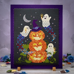 May include: A cross-stitch embroidery design featuring a stack of three jack-o'-lantern pumpkins with a witch's hat on top. Three ghosts with glowing eyes are flying around the pumpkins. The design is set against a dark background with stars.