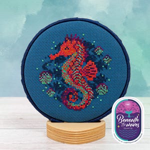 May include: A cross-stitch embroidery featuring a vibrant seahorse in shades of orange, red, and purple, surrounded by green and blue accents. The design is set on a dark blue fabric, mounted in a round frame, and displayed on a wooden stand.