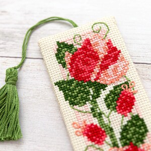 Pode incluir: Marcador de livros bordado em ponto cruz com uma rosa vermelha e rosa, folhas verdes e uma borla verde. O marcador retangular tem um fundo creme, mostrando o design floral detalhado.