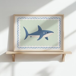 May include: A framed print of a cartoon shark in blue and white. The shark is set against a pale green background with a scalloped border, within a wooden frame. The artwork is displayed on a wooden shelf, adding a touch of coastal charm.