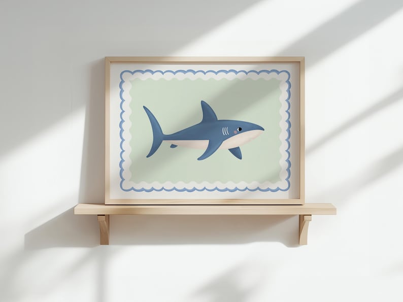 May include: A framed print of a cartoon shark in blue and white. The shark is set against a pale green background with a scalloped border, within a wooden frame. The artwork is displayed on a wooden shelf, adding a touch of coastal charm.