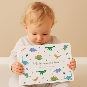May include: A baby memory book with a white cover featuring colorful dinosaur illustrations. The book has the text "Baby memory book" and the name "JAMES HENRY COLLINS" printed on the cover. The baby is holding the book.
