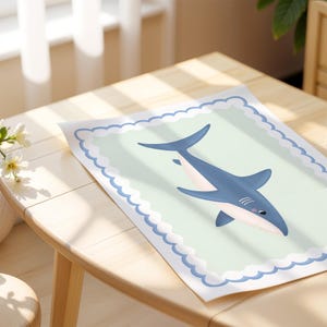 May include: A framed print with a cartoon shark illustration. The shark is blue and white, set against a pale green background with a scalloped border. The artwork is displayed on a wooden table.