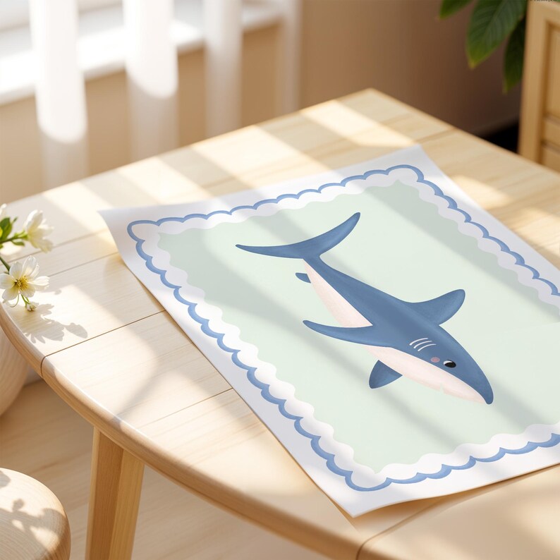 May include: A framed print with a cartoon shark illustration. The shark is blue and white, set against a pale green background with a scalloped border. The artwork is displayed on a wooden table.