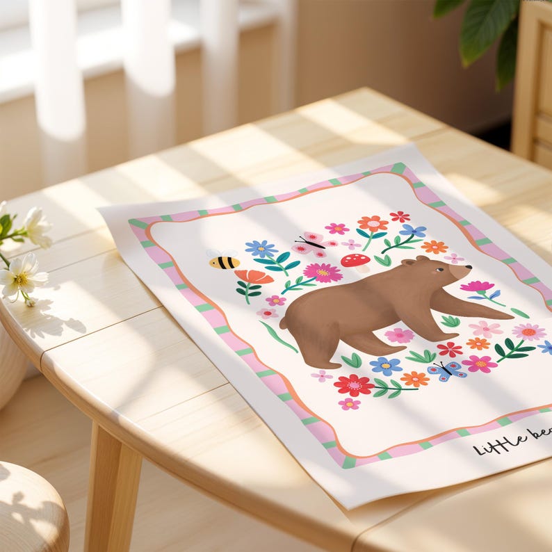 May include: A whimsical art print featuring a brown bear surrounded by colorful flowers, butterflies, and a bee. The artwork has a pink and green striped border and the words "Little bear" in the bottom right corner. The print is on a light cream background.