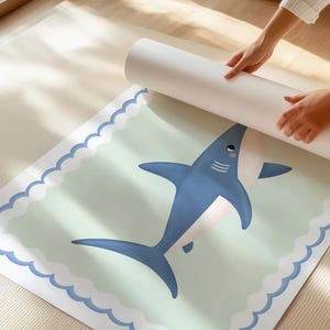 May include: A rolled print with a cartoon shark design in blue and white. The shark is on a light green background with a scalloped edge. The print is being unrolled. The image is set on a light-colored surface.