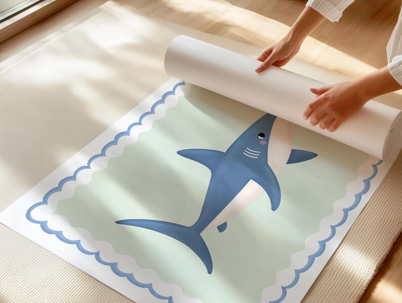 May include: A rolled print with a cartoon shark design in blue and white. The shark is on a light green background with a scalloped edge. The print is being unrolled. The image is set on a light-colored surface.
