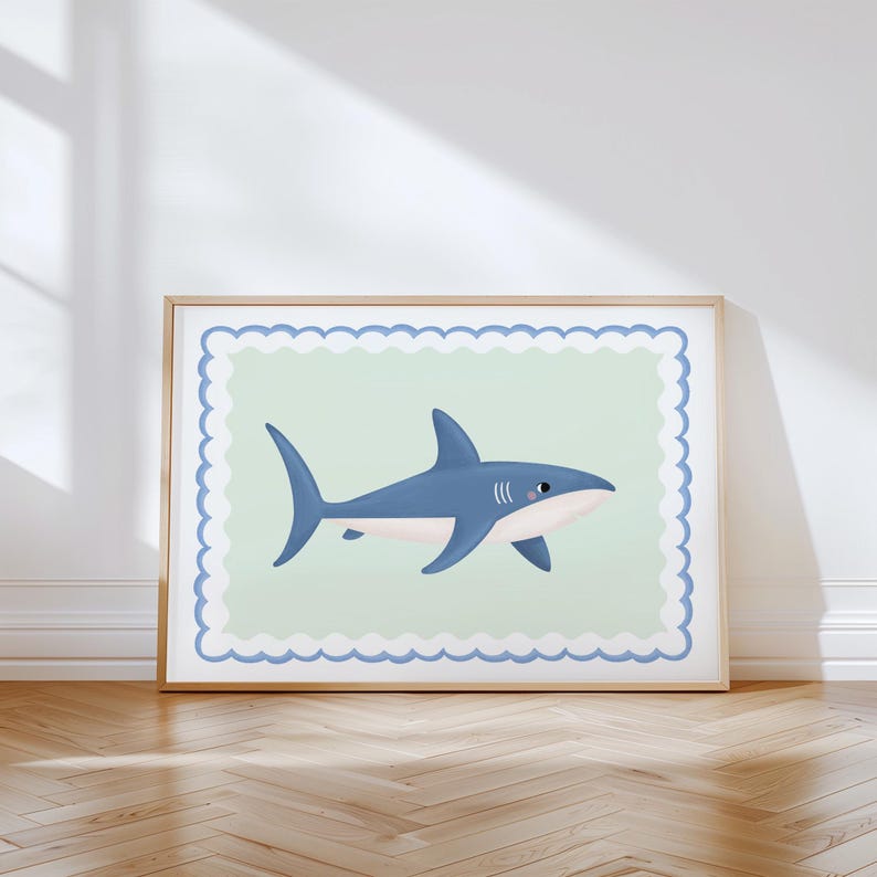 May include: A framed print with a cartoon shark illustration. The shark is blue with white details, set against a pale green background with a blue scalloped border. The frame is a light wood colour.
