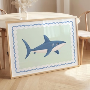 May include: A framed art print showing a cartoon shark in blue, white, and pink. The shark is set against a pale green background with a scalloped border. The artwork is presented in a wooden frame.