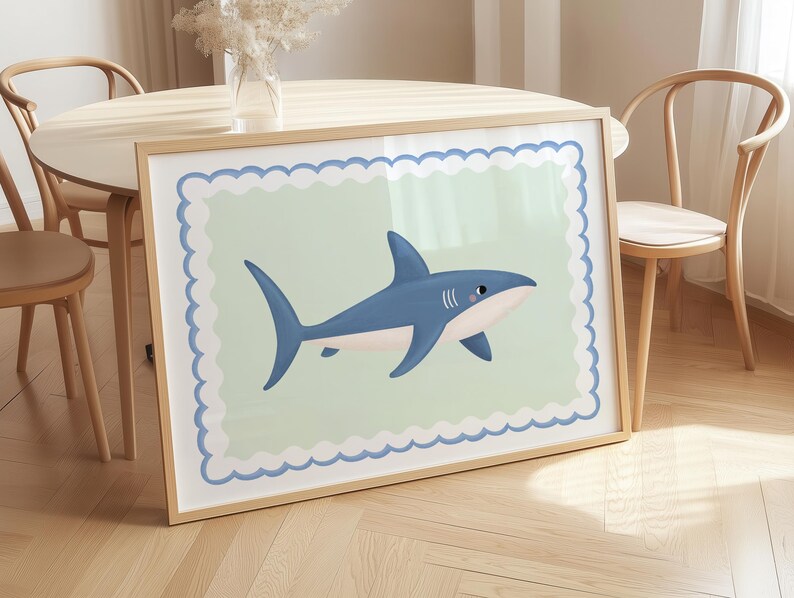 May include: A framed art print showing a cartoon shark in blue, white, and pink. The shark is set against a pale green background with a scalloped border. The artwork is presented in a wooden frame.