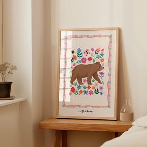 May include: A framed art print featuring a brown bear walking among colorful flowers, butterflies, and a bee. The artwork has a light wood frame and the words "Little bear" are at the bottom. The background is white with a pink and green border.