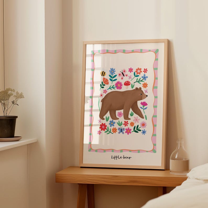 May include: A framed art print featuring a brown bear walking among colorful flowers, butterflies, and a bee. The artwork has a light wood frame and the words "Little bear" are at the bottom. The background is white with a pink and green border.