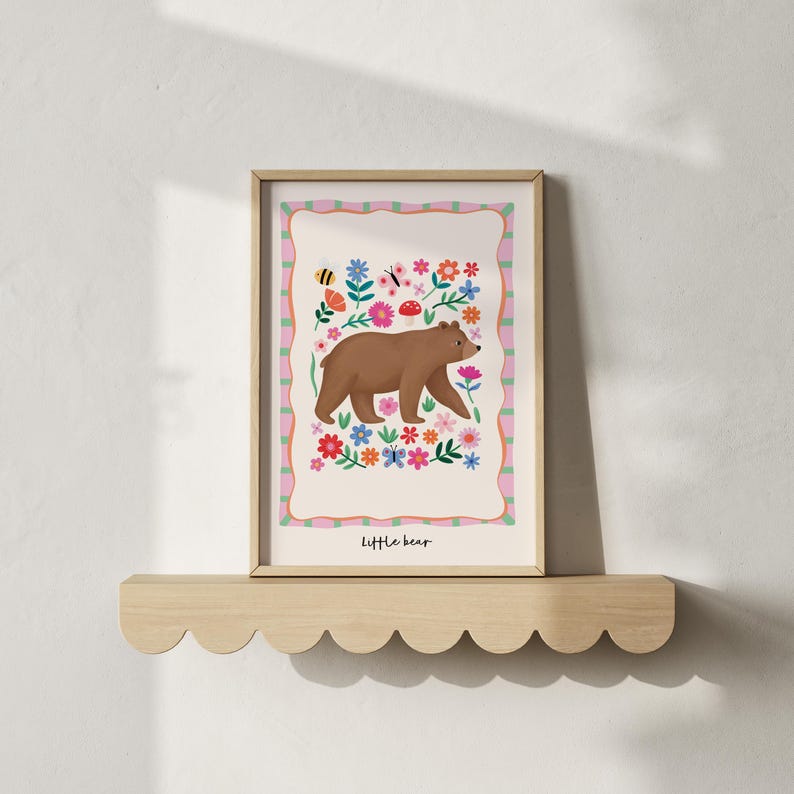 May include: A framed art print featuring a brown bear walking through a colorful floral meadow. The artwork has a light pink and green border and the words "little bear" at the bottom. The print is displayed on a wooden shelf.