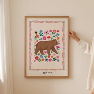 May include: A framed art print featuring a brown bear walking among colorful flowers, butterflies, and a bee. The artwork has a light beige background with a pink and green border. The words "Little bear" are at the bottom.