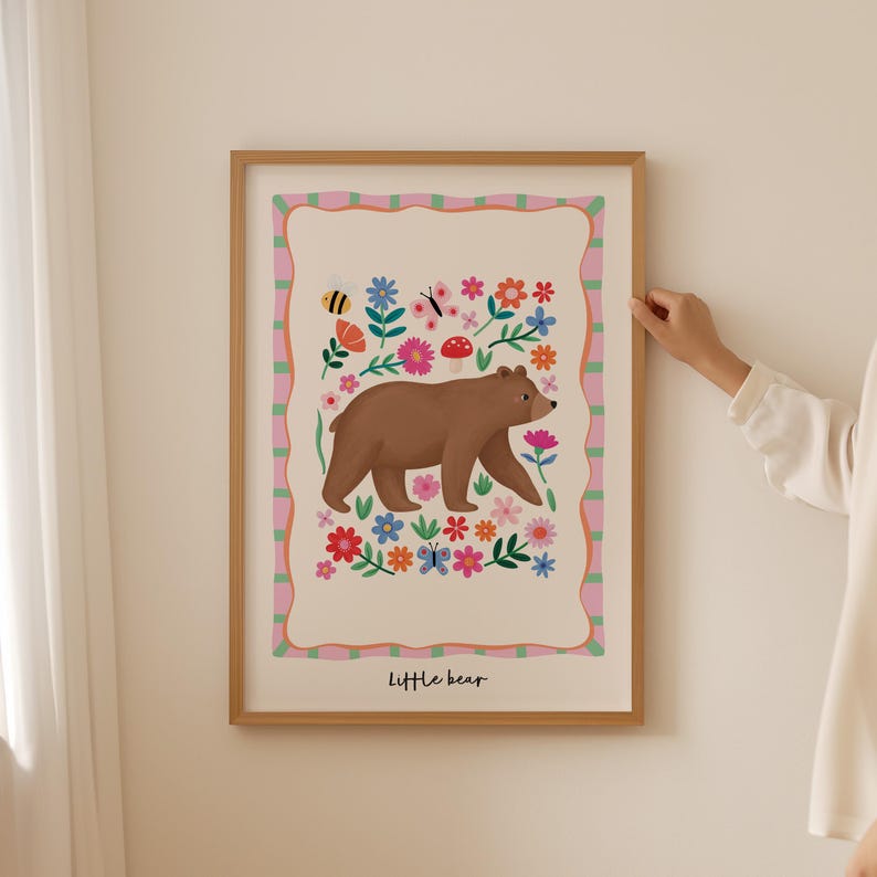 May include: A framed art print featuring a brown bear walking among colorful flowers, butterflies, and a bee. The artwork has a light beige background with a pink and green border. The words "Little bear" are at the bottom.